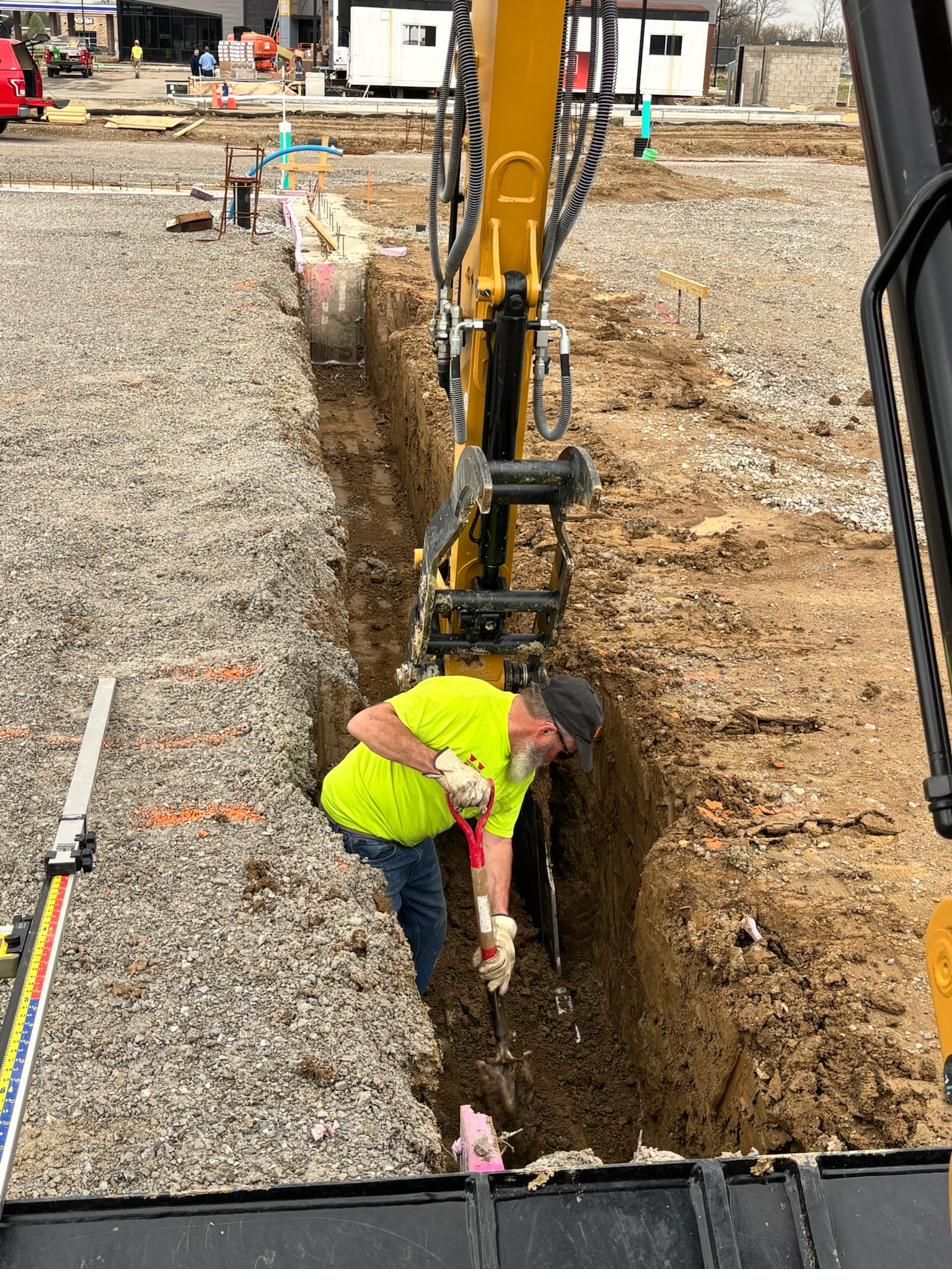Crew member checking grade in a footing trench beside the excavator stick